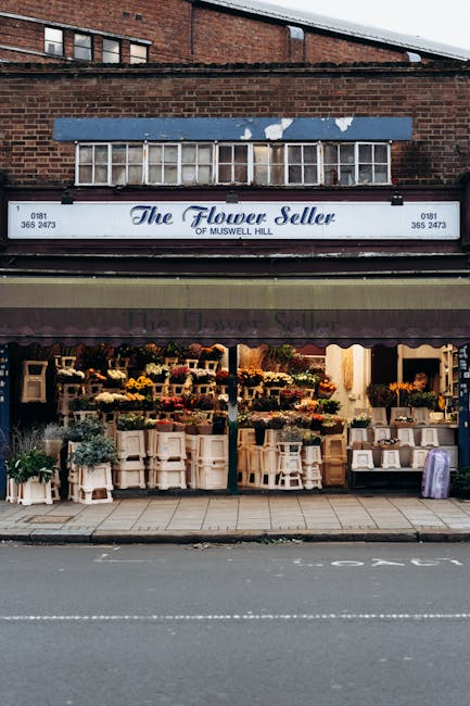 The exterior of 'The Flower Seller' shop in Muswell Hill shows a vibrant display of fresh flowers arranged on white tiered shelves and wooden crates in the storefront window. The flowers include color