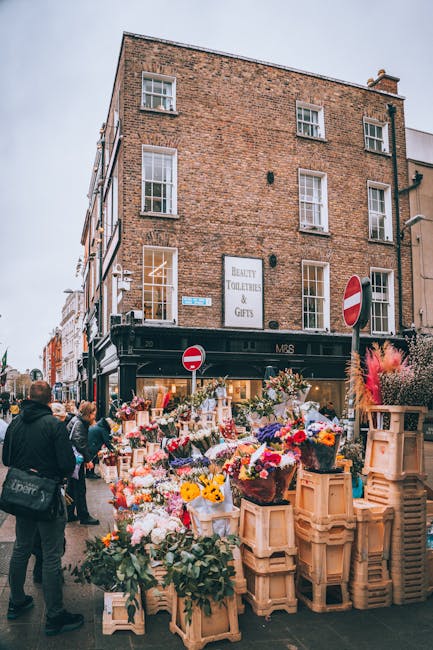 An outdoor street scene featuring a flower stall set up in front of a brick building with a black storefront. The stall displays a vibrant array of fresh flowers, including roses, tulips, sunflowers,